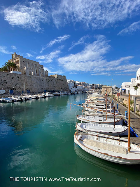 Tiny white fishing boats moored in a harbour lined by white cottages, and a sandstone castle behind the massive high sandstone harbour wall under a blue sky.