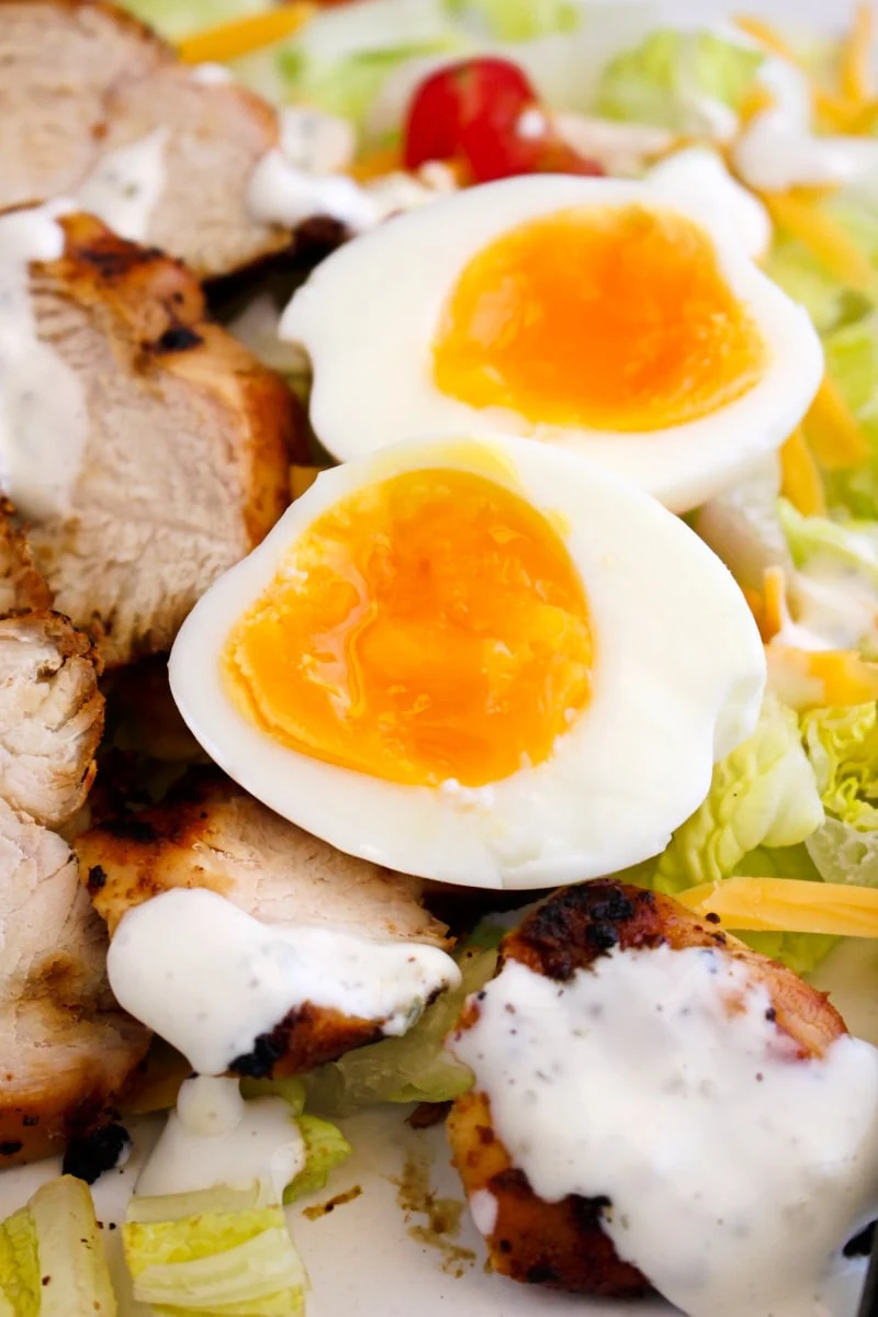 Two air fryer soft boiled eggs on a plate with lettuce, tomatoes, and sliced grilled chicken breast drizzled with ranch dressing.