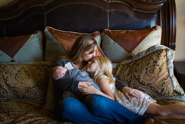 Mom, holding both children in her arms, takes a moment to give her oldest child a kiss on her forehead.  The child has her face tilted up, smiling, in response to receiving this loving gesture.  In the other arm, Mom is holding their newest family member, who is only 3-weeks old.  Newborn Session was held in my client's home located in Oklahoma City, OK.