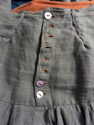 The back of a muted-teal skirt yoke, showing the rusty-orange lining where the top isn't quite aligned. The topmost and bottom two blue-iridescent shell buttons are done up, but the other five tan buttonholes are empty. Two small, angled darts are pinned into the waistband about two inches from the buttonband.