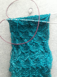 a sock on a set of circular needles.  The sock is knit in deep teal fingering-weight yarn, and has a number of small cables across the instep.  A stitch marker with a silver shell charm is clipped into the work.
