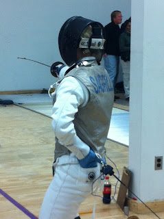 Fencer in full uniform from the back, her gloved off hand in the rear pocket of her knickers