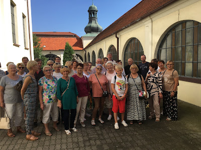 Po kcyńskim zespole pokarmelickim wycieczkę oprowadzał i przybliżył jego historię ks. proboszcz Michał Kostecki. Ksiądz Kostecki w otoczeniu zwiedzających na krużganku między Drogą Krzyżową a kościołem