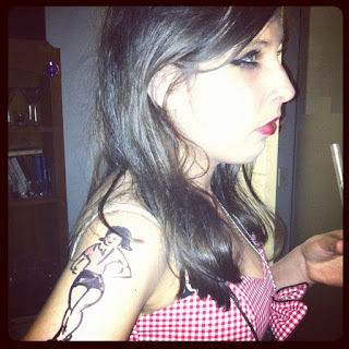 A slim, dark-haired girl in a checked dress, with a poorly drawn sharpie tattoo on her arm.