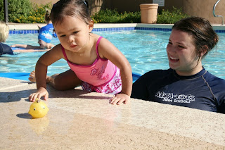 Image of a swim teacher in the pool coaching a toddler on the side of the pool after a toy duck: Toddlers Swimming Lessons