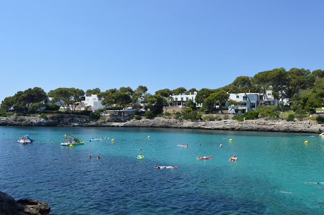 Das Foto zeigt die Cala d'Or auf Mallorca. Man sieht kristallblaues Wasser auf dem kleine Kajaks unterwegs sind, besetzt mit Menschen. Im hintergrund sind kleine Wohnhäuser zu erkennen. Wahrshceinlich handelt es sich um Ferienhäuser.