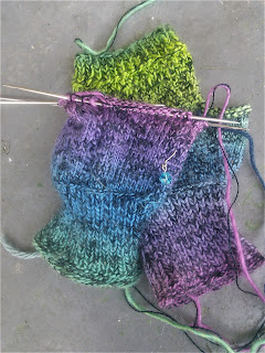 A pair of fingerless mittens, knit with yarns held double -- one yarn is black laceweight and one is a fingering-weight rainbow gradient.  One mitten is laying flat with a slipped-stitch pattern on the palm, one mitten is on double-pointed needles and laying palm down, showing stocking stitch and a small eyelet motif. 