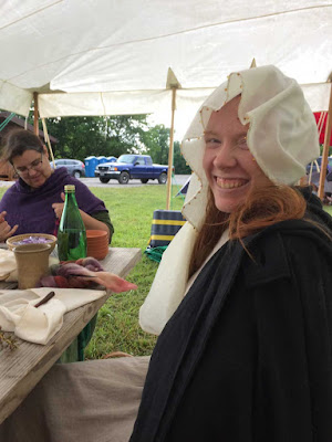 A young redheaded white woman seated at a wooden picnic bench and grinning at the camera. She's wearing a black cloak and her hair and white veil are wind-tousled. A green glass bottle, napkins and a few mugs and bowls are scattered on the table, and a white tent covers all.