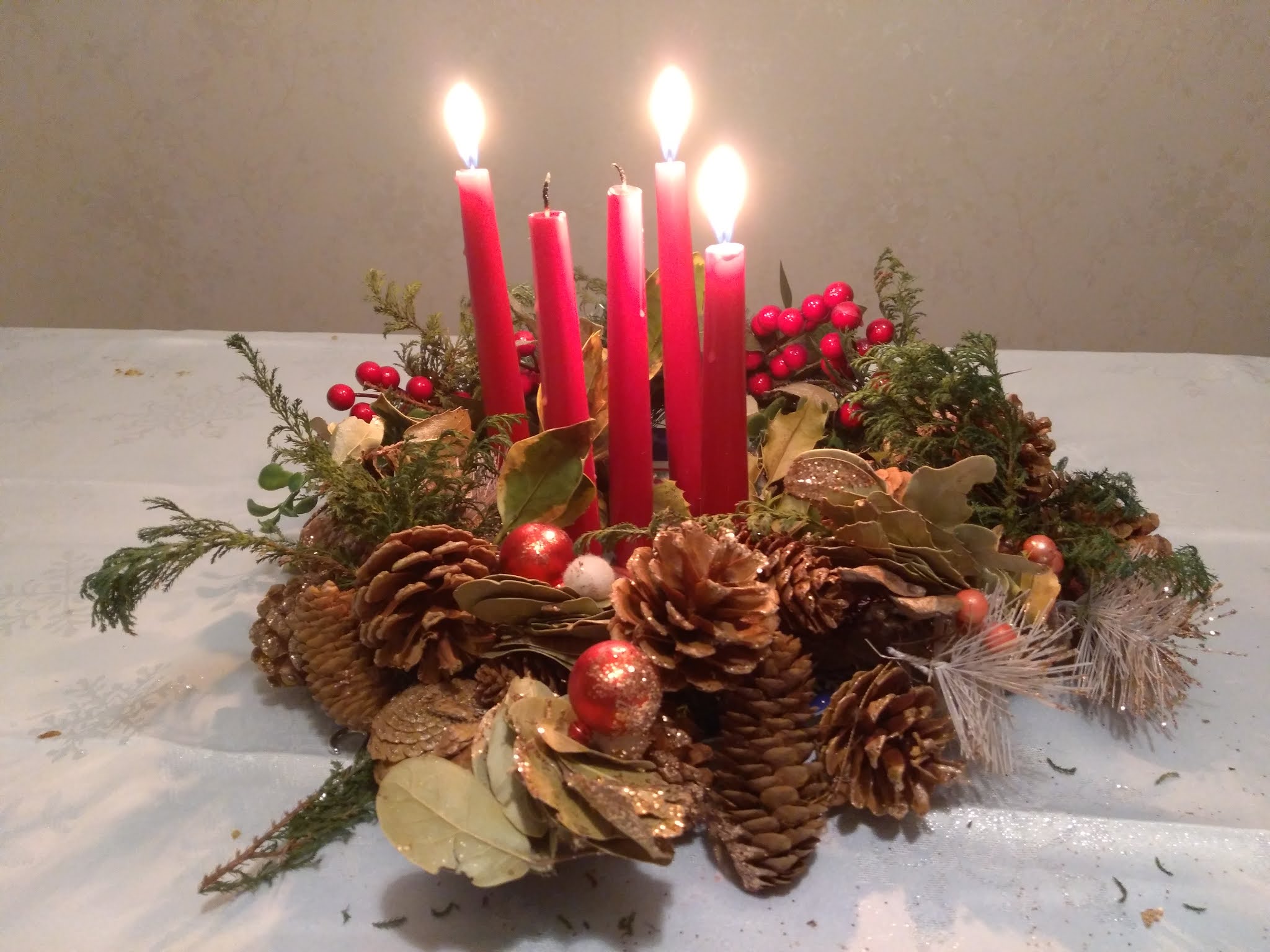 An Advent wreath on a white tablecloth, comprising holly leaves, pine cones, berries, and small fir branches. Five slim red candles are arranged in a circle in the centre, and three are lit.