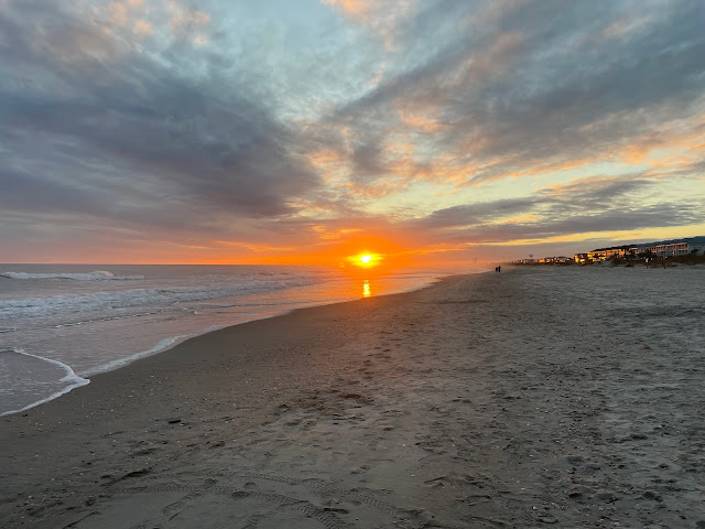 The sun setting as seen in front of the pier. The sky is full of white and gray clouds with splashes of orange, yellow, pink, and purple here and there around the sky. The water is to the left and homes and hotels are on the right.