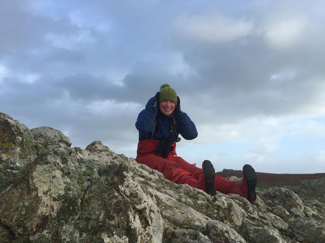 A person sat on a rock. They are holding onto their hat in the wind.