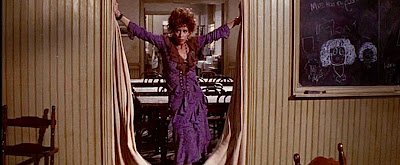 Miss Hannigan stands in a doorway, wearing a purple dress.