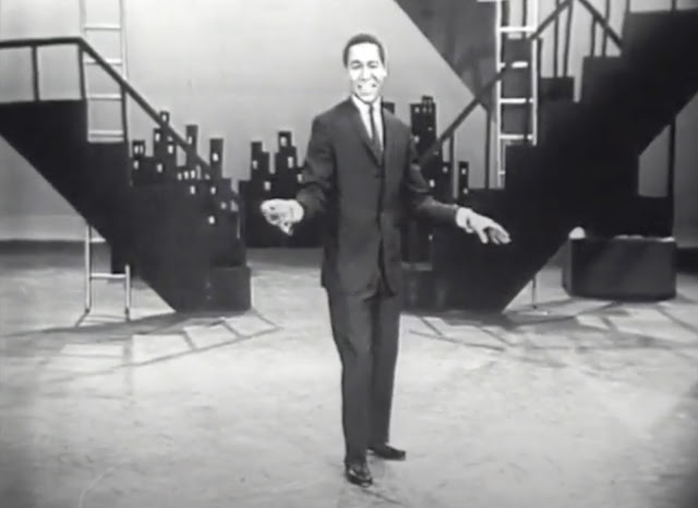 Screenshot from a 1960 TV show where Arthur Duncan signs and tap dances in front of a set design mimicking a city skyline.