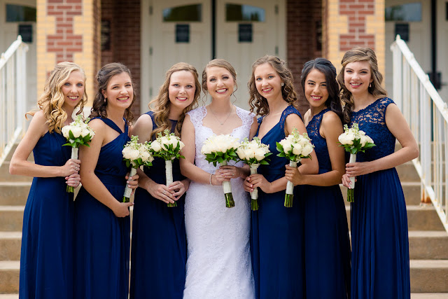 On the morning of Saturday, July 8th, the rainclouds were threatening to unleash their fury, but it didn’t matter because this was Anna and Ben’s wedding day, and for that reason alone, this day would sparkle! From the moment we got to the church, activities were in full gear…fun and impromptu pictures of the bride with her bridesmaids, as they assisted Anna  into a wedding dress that was seemingly made for her…a mermaid-shaped, intricately laced, floor length, white  gown, with a scalloped V-neck front, and a diamond-shaped, cut-out in the back.  An embroidered handkerchief was tucked inside her garter as a “remembrance” of her grandmother.  The six bridesmaids wore individually designed, royal blue chiffon gowns, and each carried a similar version of Anna’s lush bouquet of pale, ivory roses and white lilies.    Impromptu shots of the relaxed and charismatic groom with his groomsmen came next.  The comradery they share was evident in every photograph!  Then it was time to squeeze in a sentimental “first look” for the bride’s dad before the romantic “first look” with the groom, and you can tell from the expression on Ben’s face…and the height of his jump…how excited he was to finally see her!     First Church OKC was an ideal location for this couple’s wedding.  Located in the heart of Oklahoma City, its ministry began during the land run of 1889, and has continued to share the gospel through two wars and the infamous April 1995 bombing.  The large, stained glass windows, dark mahogany wood, and simple, yet elegant sanctuary was the perfect backdrop for these two young people who were meant for each other.     When asked what the best part of the day was, Anna said, “getting to see Ben for the first look, and his reaction to seeing me.”  Ben’s response was “getting to see Anna walk down the aisle and say our vows to each other.”     Anna felt that her wedding day went smoothly, but if she were to do it over again, she would hire a wedding planner and a florist to take some of the responsibility off herself, family, and friends.  Her favorite piece of advice to a prospective bride and groom:  “When things get stressful or overwhelming—and at some point they will—just take a break, step back, and enjoy.”    After the couple exchanged vows, guests were invited to participate in a fun, romantic, and lively reception that included the “first dance,” dances with the parents, toasts to the bride and groom, a sit-down dinner, the cake cutting, throwing of the bouquet and garter…and lots and lots of dancing!  At the end of the evening, it was time for friends and family to gather together and wish this fun-loving couple farewell and best wishes for a wonderful life together!  