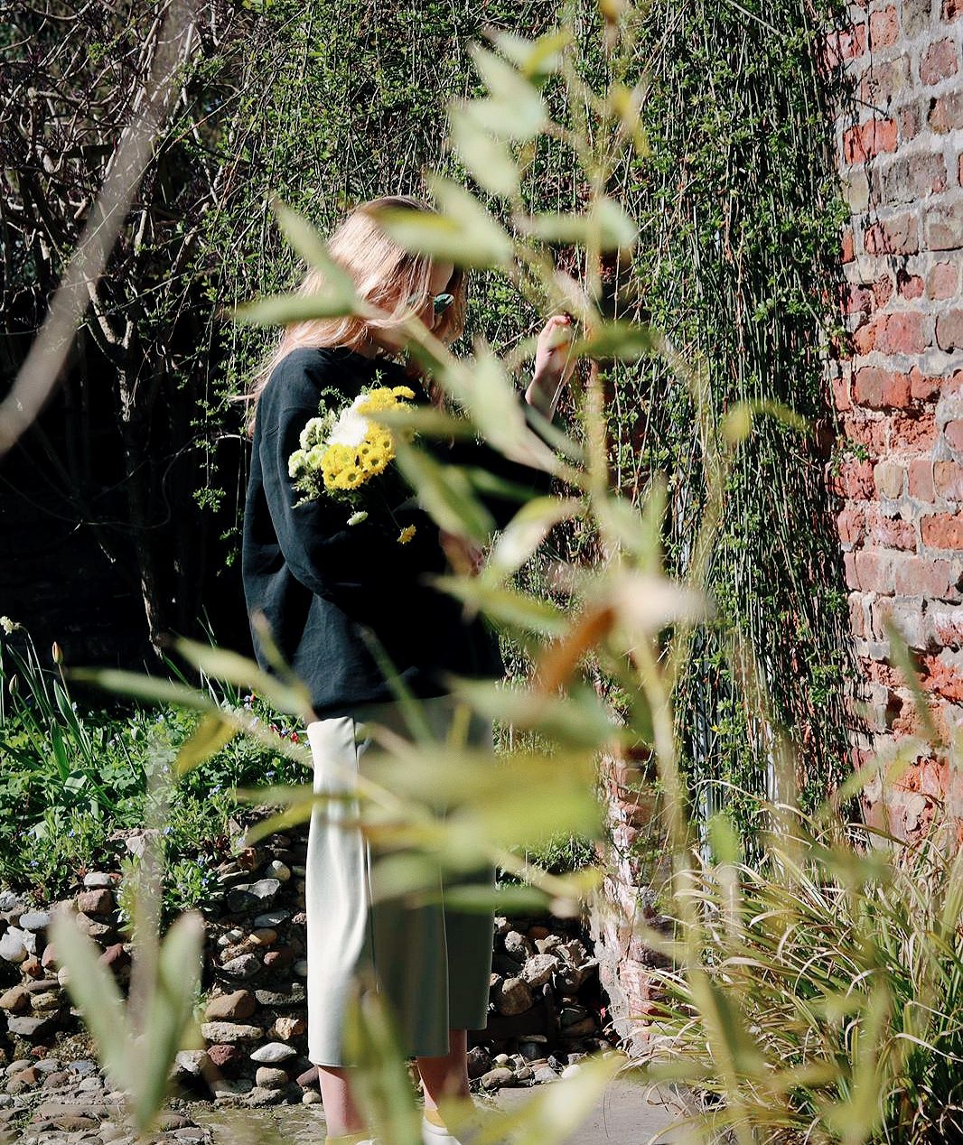 Amy is standing in an overgrown garden, hidden behind some greenery, holding a bunch of flowers in one arm