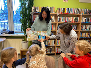 Pani bibliotekarka trzyma w rękach otwartą książkę, druga pani trzyma komórkę przed tą książką. Dziewczynka dotyka palcem ekranu telefonu. Tło regały z książkami