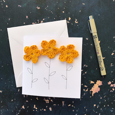 3 crocheted yellow flowers stuck on a greetings card. Stems have been drawn in black pen, which sits to the right hand side of the card