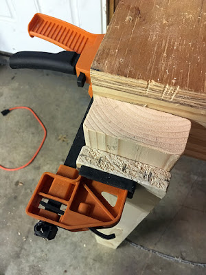 A shot from above showing the end of a glued-up pair of boards, clamped sideways with a black-and-orange clamp to a wooden platform lip, with three small marks in a line across the end of the shorter, outer 1x4 board.