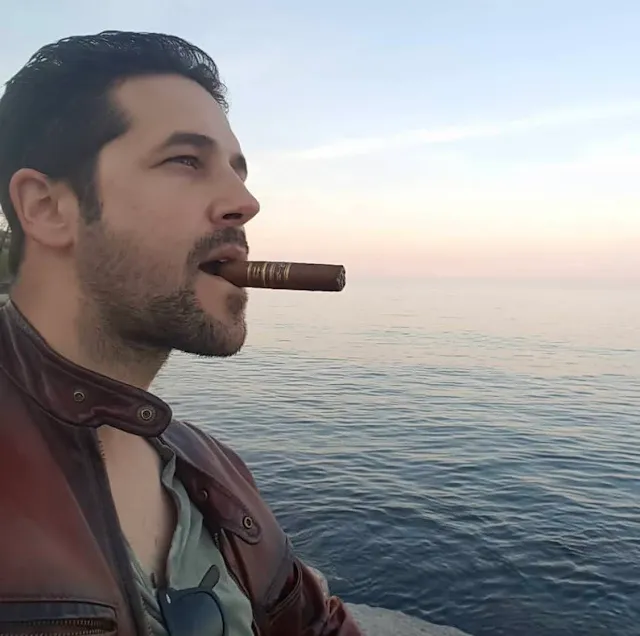 Dark-haired while manicured wearing red leather jacket and smoking a cigar with a beautiful dusk in the background while manicured wearing red leather jacket and smoking a cigar with a beautiful dusk in the background