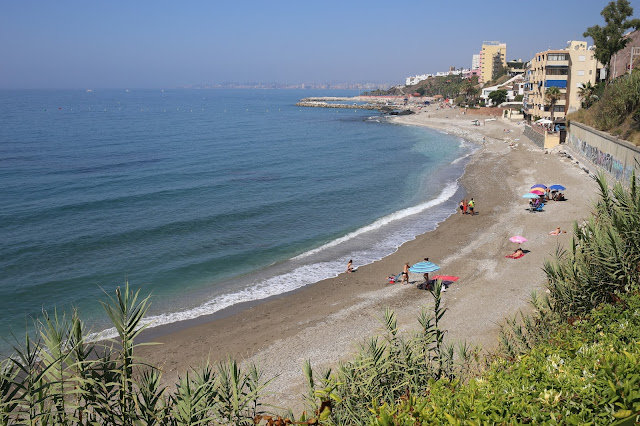 Playa con las azules aguas del mar, acantilados con vegetación y edificios a su espalda.