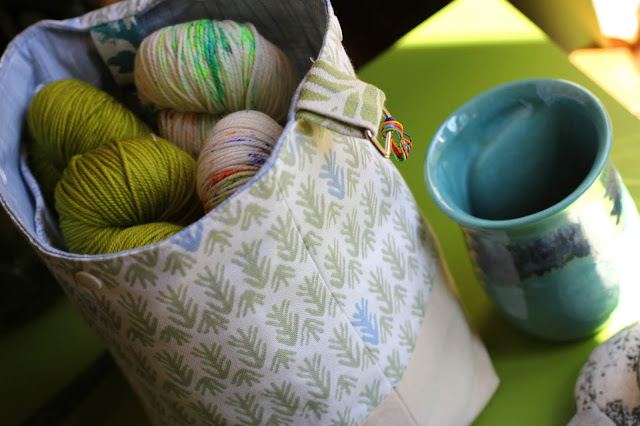 5 hanks of yarn fit inside a project bag that has a sage patterned exterior and blue chambray lining interior. Two fabric loops made of meaningful scrap hold a swivel snaphook as a yarn guide and a ring for stitch markers. Handwarmer mug with late afternoon lukewarm coffee sits next to the bag.