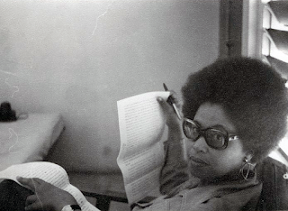 Black and white image of Sylvia Wynter, unknown date. She has an afro hairstyle and is wearing very large earring and sunglasses. She is leaning back in a chair and looking up at the camera. She is holding type written document and a pen.