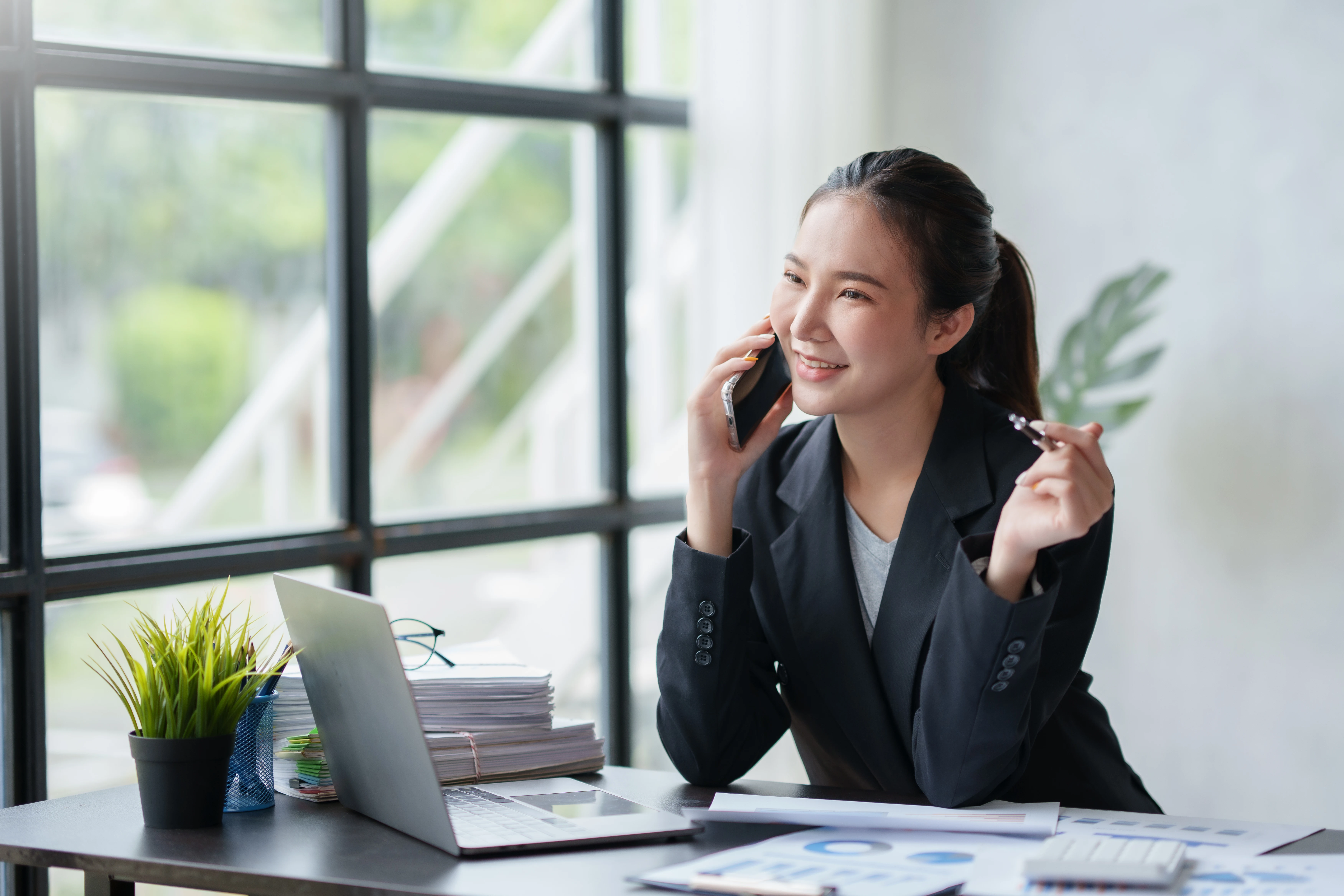 Empresária asiática sorridente aproveitando o telefone, respondendo perguntas, conversando com clientes ao telefone e comentando no escritório.