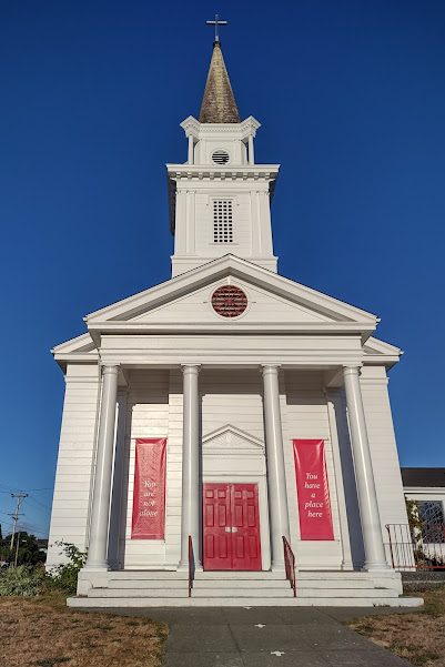 Photo of a white-pained wooden church with a pointy spire and multiple columns, against a deep blue sky. The curch has a red door and some red banners hanging on its porch.