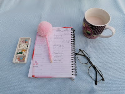 Auf hellblauem Stoff liegen folgende Gegenstände von links nach rechts:eine gefüllte weiße Tablettenbox, ein aufgeschlagener Termnplaner, darauf ein rosa Stift mit rosa Puschel am Ende, eine Tasse Kakao und darunter eine Brille.