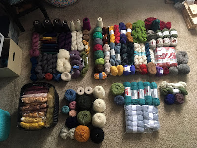 An array of yarn laid out on a beige carpeted floor, mostly in twisted hanks, but some in hand-would cakes and some on cones. There are six distinct sections, from a square at upper left to a large rectangle at upper right, with three smaller sections below, and a small black plastic tote crammed full of clear bags holding gleaming yellow, brown, tan, and red silks. Overall there's a lot of blue and green, but quite a bit of white, grey, and pink-red, too.
