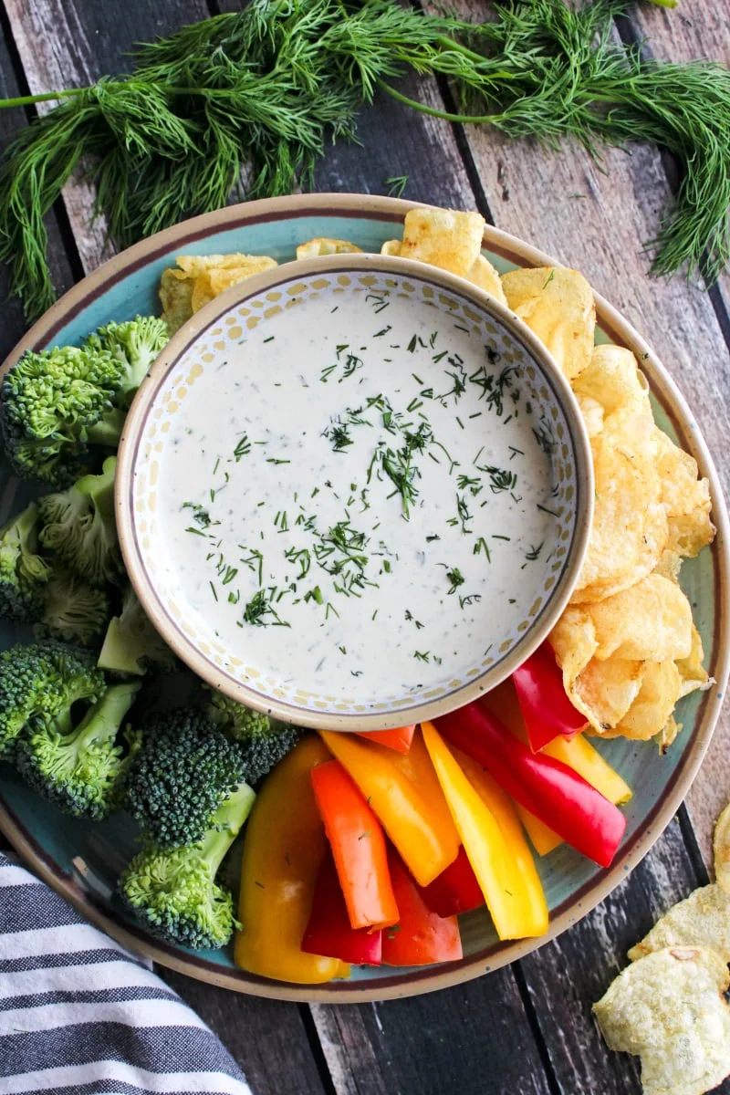 Top view of Dill Dip in a tan bowl on a blue plate surrounded by veggies and chips.