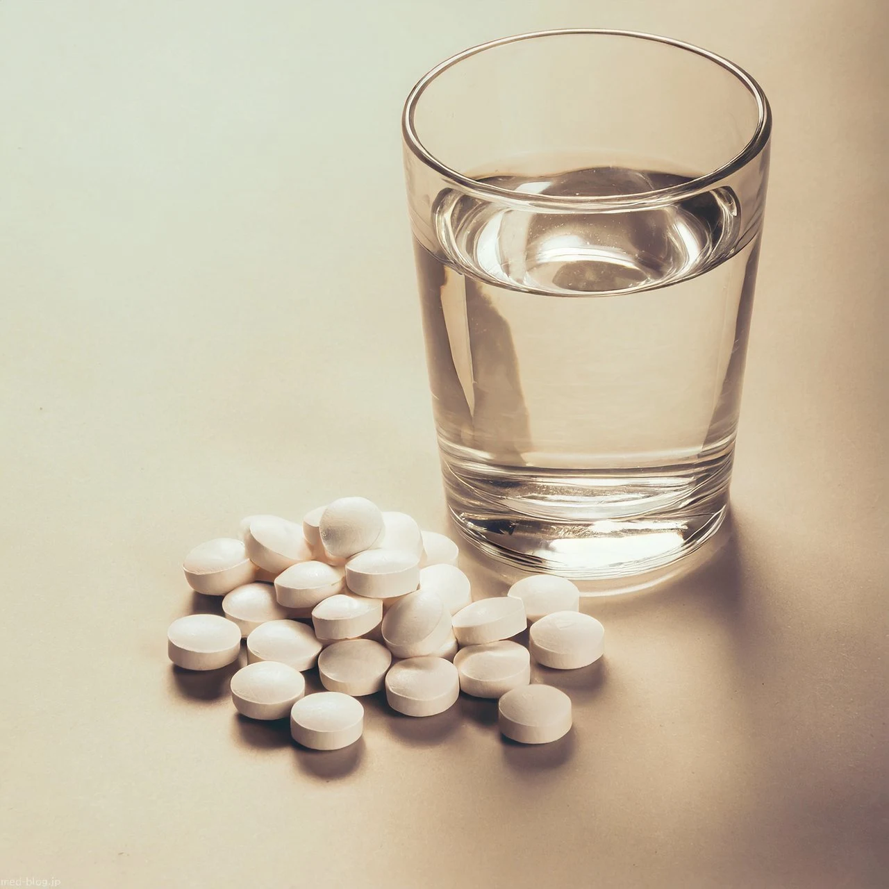 白い机の上に20条ほどの白い錠剤と水の入った透明なコップが置かれている写真.jpg (1280×1280)