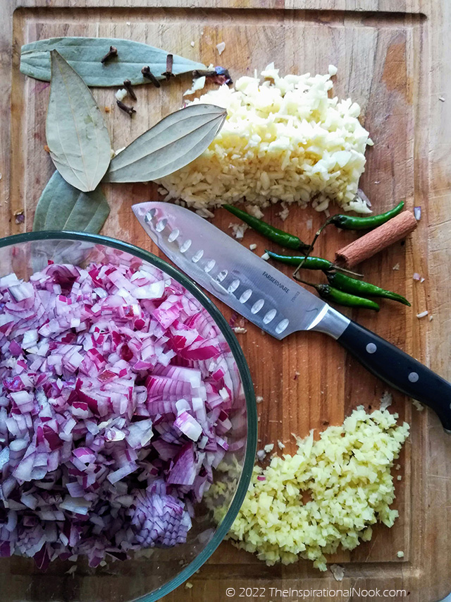 Ingredients for bafad pork, red onions, bay leaves, cloves, garlic, ginger on a cutting board with a santoku knife