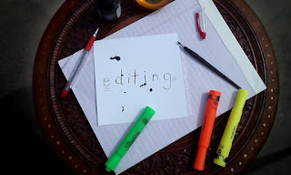 Top down view of the top of a decorative circular table topped with a purple legal pad, a red pen, a bottle of black india ink, highlighters, a dip pen, and a square of white card stock with editing writen on it.  Ink splatters across the card stock and there are grammatical correction symbols in red ink.  