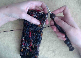 A crochet mitten cuff done in a textured crochet stitch.  It uses fingering-weight variegated yarn in yellow, orange, red, and black.  Two silver stitch markers are clipped into the cuff.