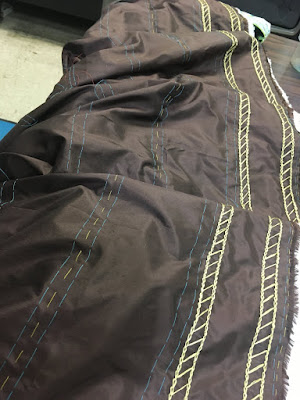 A long piece of dark brown silk, laid out in loose folds along a table, with turquoise and yellow basted guidelines running down the length, and a pair of doubled gold chain stitch lines, each with closely spaced diagonals between them.