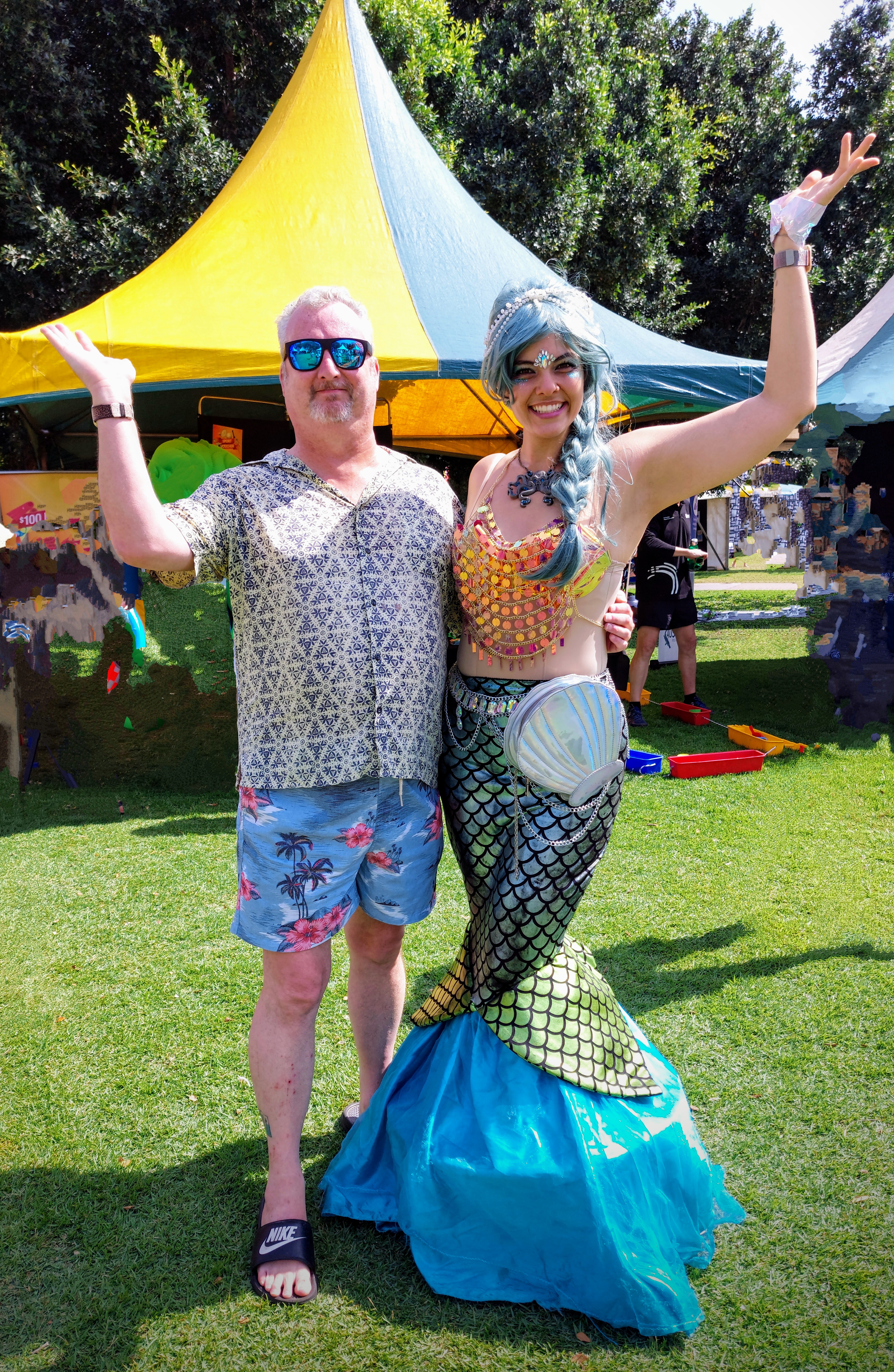 In front of a fair tent Mike stands with his arm extended next to a woman dressed as a mermaid as she does the same. It's a sunny Darwin day and everyone is smiling.