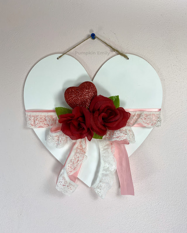 A white heart wall hanging with roses, a glitter heart, leaves, lace, and ribbon.