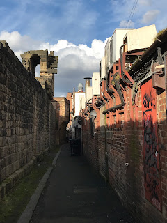 A view of a path that runs at the back of the town wall.  On one side is the old town wall, there is a tarmac path in the middle and then on the other side the red brick walls and air conditioning units of the various industrial buildings.  Photo by Kevin Nosferatu for the Skulferatu Project.