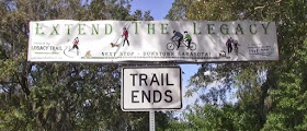 Two signs hanging on trees say trail ends and extend the Legacy Trail sign in Sarasota, Florida.