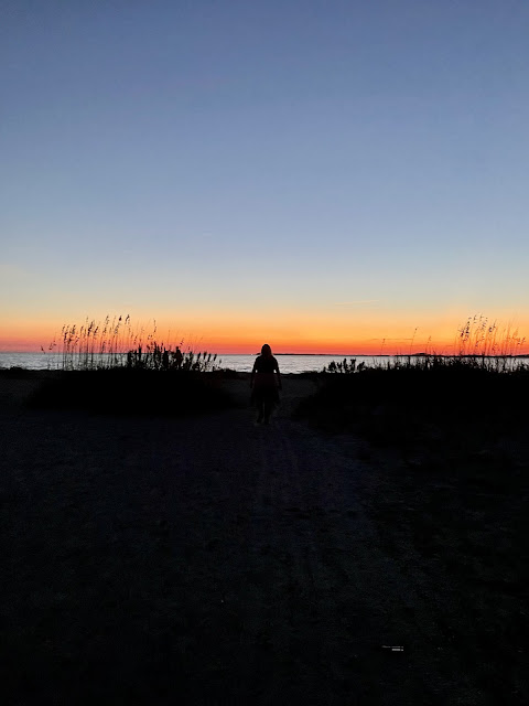 Me walking on the beach path to the beach. I am a black silhouette because of the time of day it is. Because of am walking toward the beach the photo captures me from behind. There is beach grass to the left and right of me with the darkened sky above. There is still an orangeish hue on the horizon from where the sun had just set. The ocean is before me.