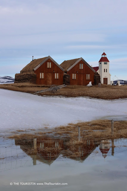 The reflection of a white red-roofed church and two wooden farmhouses in flooded snow-covered fields, under a light blue early evening sky.