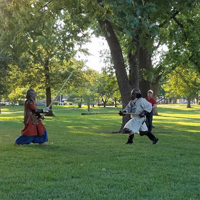 Two people in medieval armor and clothing, facing each other in a green, sunlit park. The person on the left is holding a greatsword and wearing a long, brown leather armor coat over a longer red coat and very full blue pants. The person on the right is in a long white tunic and black pants, and is moving toward the person in red with their spear levelled. A man in a red polo and jeans is just visible behind the person in white.