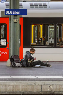 Einer mit Gitarre sitzt am Bahnhof auf dem Boden und singt Lieder aus einem mitgebrachten Gesangbuch.