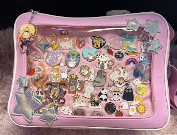 A pink ita bag that has a clear window so you can see the pins that are on displayed. The pins are all different and varies from encouragement words to anime pins