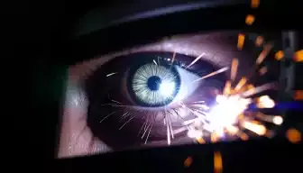 A close-up of an eye with a bright, intense glare reflecting arc welding sparks, showcasing a protective welding face shield partially obscured, surrounded by dark shadows and glowing embers in the background, emphasizing the contrast between the eye's vulnerability and the hazardous environment.