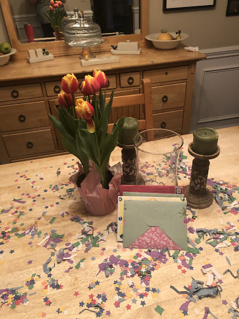 The dining room table with  table confetti, an empty vase, two beige candlestick holders with green candles have burned down on them, a tulip plant, and my birthday cards on it.