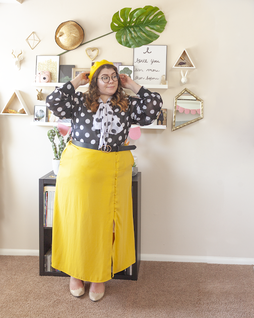 An outfit consisting of a yellow beret, a white on black polka dot blouse with a black on white polka dot neck tie attached, tied in a bow, tucked into a yellow button down midi skirt and beige slingback heels.