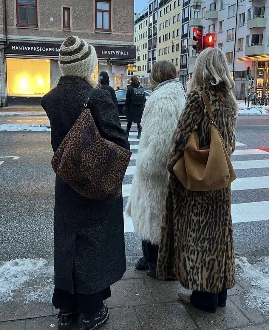 Three women with blonde hair wearing skull cap hat, long wool coat, long white shaggy fur coat, long leopard print fur coat, oversized hobo leather bags in tan, brown, and leopard print, black pants and boots standing at the curb at the crosswalk on the street with tall brick and grey buildings across the street