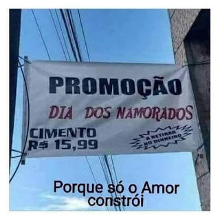 Foto em dia de céu limpo. Uma faixa publicitária retangular branca presa entre um poste e uma coluna de concreto com o texto: Promoção Dia dos Namorados; no canto inferior esquerdo lê-se: Cimento quinze reais e noventa e nove centavos; e no outro canto: A retirar no dinheiro. Abaixo, o recado: Porque só o Amor constrói. 